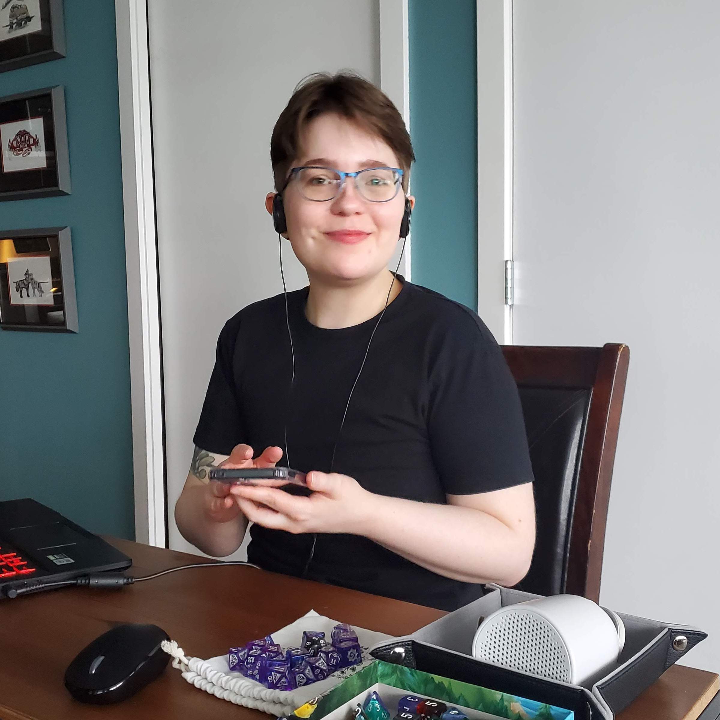 A photo of Myth, a white Metis person with short hair wearing eyeglasses and a black t-shirt, sitting in front of a laptop and some polyhedral dice